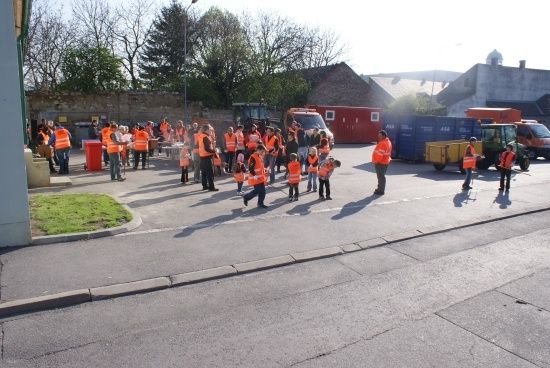 Eine Gruppe von Menschen in orangefarbenen Westen versammelt sich auf einem Parkplatz mit mehreren Fahrzeugen und einem Gebäude im Hintergrund.