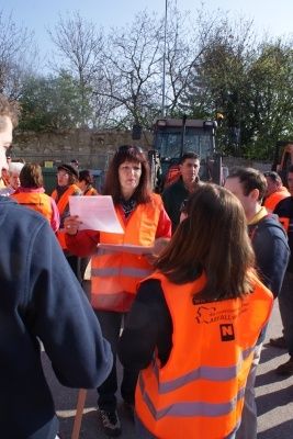Eine Gruppe von Menschen in orangefarbenen Warnwesten steht draußen, einer hält ein Papier und spricht mit anderen. Ein Baufahrzeug ist im Hintergrund zu sehen.