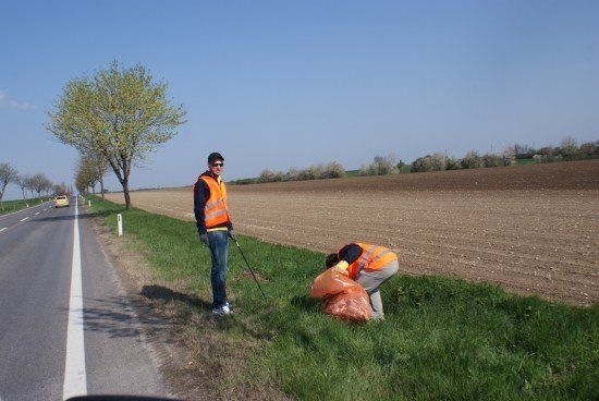 Zwei Personen in orange Westen sammeln Müll am Straßenrand. Eine beugt sich, die andere steht mit einem Stock.