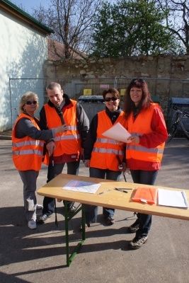 Vier Personen in orange Sicherheitswesten stehen um einen Tisch mit Papieren in einer Außenumgebung.