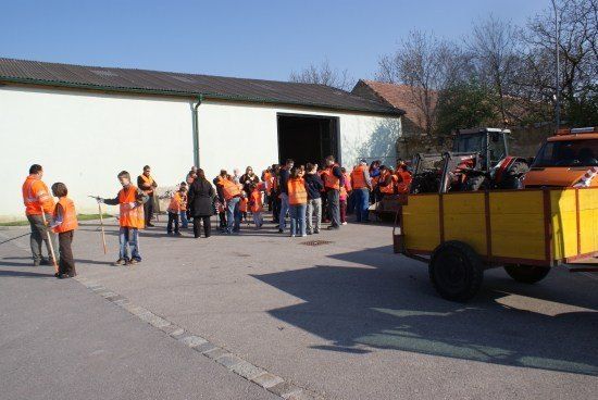 Eine Gruppe von Menschen in orangefarbenen Westen steht vor einem Gebäude mit einem gelben Anhänger in der Nähe. Sie scheinen auf Anweisungen zu warten.
