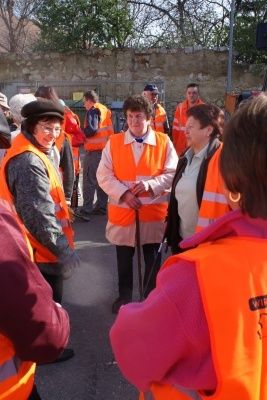 Eine Gruppe von Menschen in orangefarbenen Westen steht und unterhält sich im Freien. Eine Frau in der Mitte hält einen Stock. Hinter ihnen befindet sich eine Mauer mit einem Metallzaun und Bäumen.