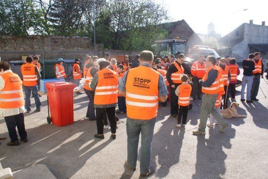 Eine Gruppe von Menschen in orangefarbenen Westen steht in einer Reihe, möglicherweise an einer Gemeinde-Aufräumaktion beteiligt. Ein roter Mülleimer steht links, und ein Maschinenfahrzeug ist dahinter geparkt.