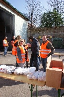 Eine Gruppe von Menschen in orangefarbenen Sicherheitswesten steht um einen Tisch mit verschiedenen Gegenständen darauf, möglicherweise in Vorbereitung auf eine Veranstaltung.