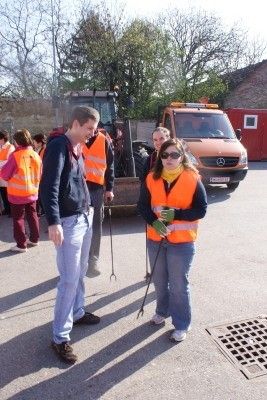 Eine Gruppe von Menschen in orangenen Westen steht auf einem Parkplatz, mit einem Traktor und einem LKW im Hintergrund. Ein Mann und eine Frau stehen sich gegenüber und halten beide Stöcke.