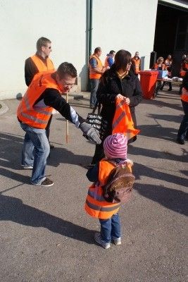 Ein Mann in einer orangefarbenen Warnweste gibt einem kleinen Kind, das eine rosa Mütze und einen Rucksack trägt, eine orange Flagge. Dahinter stehen mehrere Personen in orangefarbenen Westen in der Nähe eines Gebäudes.