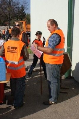 Ein Mann in einer orangefarbenen Sicherheitsweste liest ein Dokument, während zwei Kinder in orangefarbenen Westen neben ihm stehen. Eine Frau geht hinter ihnen, und ein Lastwagen ist im Hintergrund geparkt.