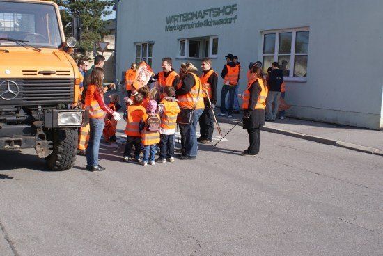Eine Gruppe von Menschen in orangefarbenen Westen versammelt sich vor einem Gebäude mit einem Schild, auf dem Wirtschaftshop steht. Ein großer orangefarbener LKW ist in der Nähe geparkt.