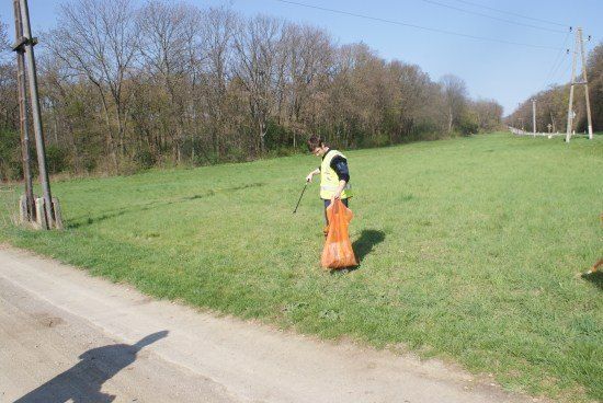 Eine Person in einer gelben Weste sammelt Müll in einem Grasfeld in der Nähe einer unbefestigten Straße.