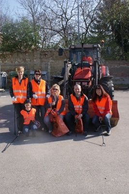 Eine Gruppe von Freiwilligen in Warnwesten posiert für ein Foto vor einem Traktor. Sie halten Müllsäcke und Stöcke in den Händen. Im Hintergrund sind eine Mauer und Bäume zu sehen.