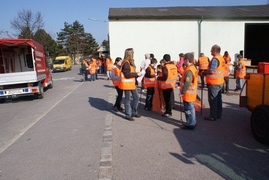 Eine Gruppe von Menschen in orangefarbenen Westen steht auf einem Parkplatz, einige halten Papiere. Ein roter LKW ist in der Nähe geparkt. Ein Gebäude mit einer großen offenen Tür befindet sich im Hintergrund.