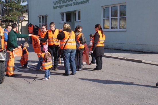 Eine Gruppe von Menschen in orangenen Westen, einschließlich Kindern, versammelt sich auf einer Straße, möglicherweise für eine Gemeinschaftsveranstaltung. Im Hintergrund ist ein Gebäude mit der Aufschrift 'Wirtschaftsschule Schwadorf' zu sehen.