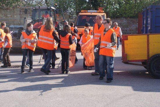 Eine Gruppe von Menschen in orangefarbenen Westen steht auf einem Parkplatz, möglicherweise an einer Müllsammelaktion beteiligt. Sie halten Plastiktüten und scheinen Müll zu sammeln. Im Hintergrund befinden sich zwei Fahrzeuge, eines davon ist ein Traktor.