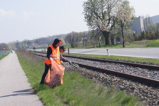 Zwei Personen in orange Weste sammeln Müll in einem Grasbereich neben einer Bahnlinie.