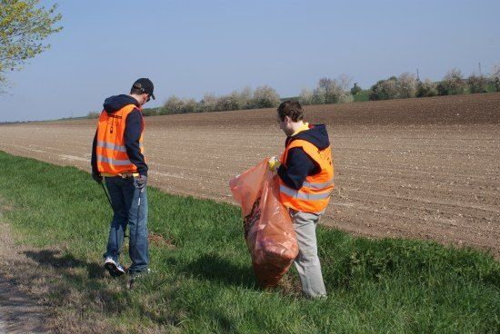 Zwei Personen in orangen Westen sammeln Müll in einem Grasbereich in der Nähe eines gepflügten Feldes unter einem klaren Himmel.