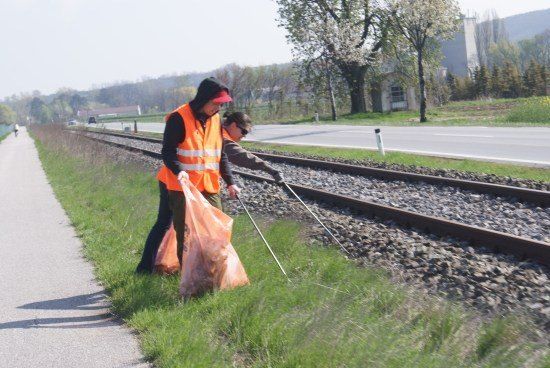 Zwei Personen in Warnwesten sammeln Müll entlang einer Bahnstrecke ein.