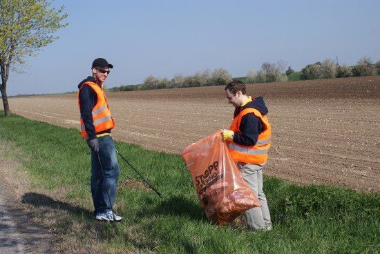 Zwei Männer in orange Weste sammeln Müll in einem Grasbereich in der Nähe eines Feldes. Einer hält einen Müllsack, während der andere einen Stock benutzt.
