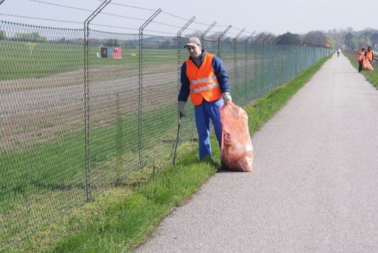 Ein Mann in einer orangefarbenen Warnweste sammelt Müll entlang eines Weges in der Nähe eines Zauns.