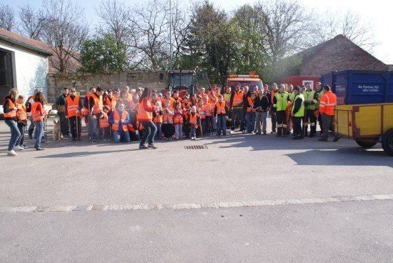 Eine Gruppe von Menschen in orangefarbenen Westen steht auf einem Parkplatz mit einigen Fahrzeugen im Hintergrund.