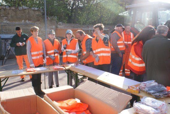 Eine Gruppe von Menschen in orangenen Westen versammelt sich um einen Tisch, möglicherweise in Vorbereitung auf eine Veranstaltung oder Aktivität. Einige schreiben auf Papiere, während andere zuschauen.