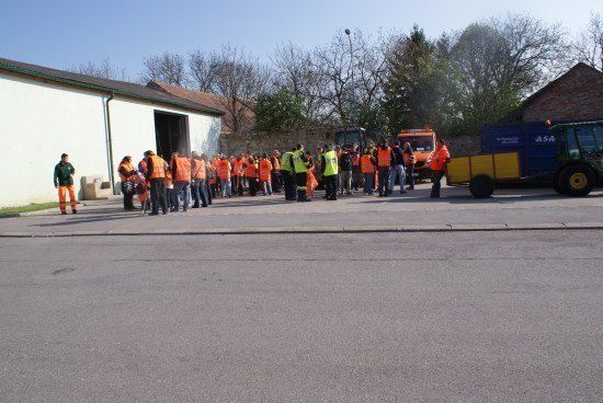 Eine Gruppe von Menschen in orangefarbenen Westen steht vor einem Gebäude, in der Nähe befindet sich ein Traktor und ein Maschinenfahrzeug.
