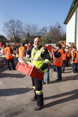 Ein Mann in einem gelben Sicherheitsweste hält eine rote Kiste voller Gemüse und steht vor einem Gebäude, mit mehreren Personen in orangefarbenen Westen im Hintergrund.