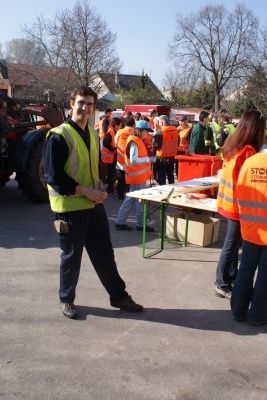 Ein Mann in einer gelben Sicherheitsweste steht auf einem Parkplatz mit einer Gruppe von Menschen in orangefarbenen Westen, in der Nähe eines Tisches mit verschiedenen Gegenständen und einem Maschinenfahrzeug im Hintergrund.