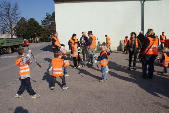 Eine Gruppe von Menschen in orangefarbenen Westen, einschließlich Kindern, versammelt sich in einem Außenbereich, möglicherweise für eine Gemeinschaftsveranstaltung. Einige halten Stöcke, und ein Lastwagen ist in der Nähe geparkt.
