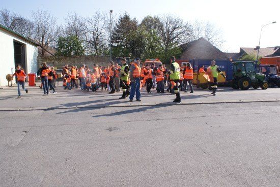 Eine Gruppe von Menschen in orangefarbenen Westen geht auf einer Straße, mit mehreren geparkten Fahrzeugen in der Nähe.