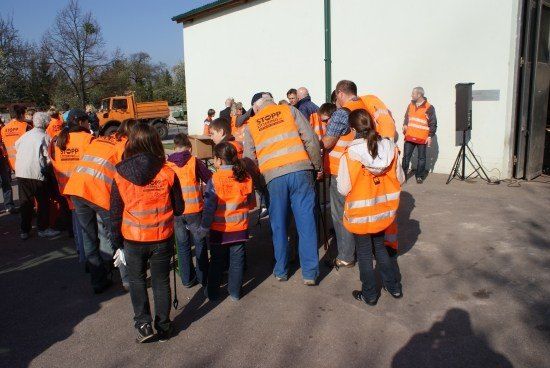 Eine Gruppe von Menschen in orange Sicherheitswesten versammelt sich vor einem Gebäude. Einige schauen einen Mann in einer grauen Weste mit dem Wort 'STOPP' darauf an. Ein Lautsprecher steht auf einem Stativ rechts.