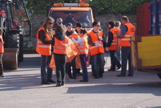 Eine Gruppe von Menschen in orangefarbenen Westen steht draußen, einige halten Werkzeuge, mit Maschinen und einer Ziegelmauer im Hintergrund.