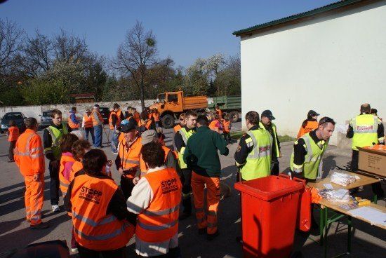 Eine Gruppe von Menschen in orangefarbenen Sicherheitswesten versammelt sich in einem Außenbereich, möglicherweise für eine Arbeitsveranstaltung. Ein roter Mülleimer und ein Tisch mit Gegenständen sind in der Nähe. Ein LKW ist im Hintergrund geparkt.