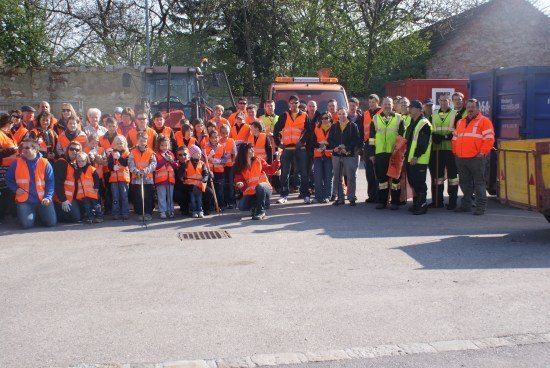 Eine Gruppe von Menschen in orangefarbenen Westen und Handschuhen posiert für ein Foto auf einer Straße mit Maschinenfahrzeugen und Müllcontainern im Hintergrund.