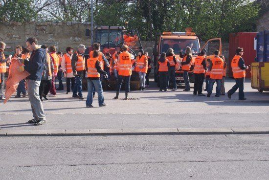Eine Gruppe von Menschen in orangefarbenen Westen steht in der Nähe einer Straße, mit Maschinenfahrzeugen und einem roten Container im Hintergrund.