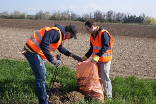 Zwei Männer in orangenen Westen sammeln Müll in einem Feld mit einem großen orangefarbenen Beutel.