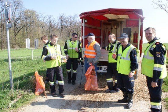 Eine Gruppe von Menschen in Warnwesten und Stiefeln steht auf einem Kiesweg, einige halten Plastiktüten in der Hand, mit einem roten Lastwagen und einem Pfosten im Hintergrund.