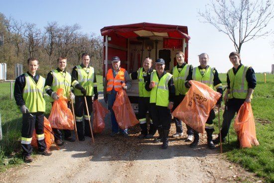 Eine Gruppe von Menschen in Sicherheitswesten steht auf einem Feld, hält orangefarbene Taschen und Stöcke. Ein roter Lastwagen ist hinter ihnen geparkt.