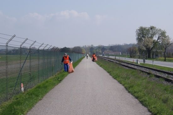 Zwei Personen in orangen Westen gehen auf einem Pfad neben einer Bahnlinie, einer trägt einen Beutel.