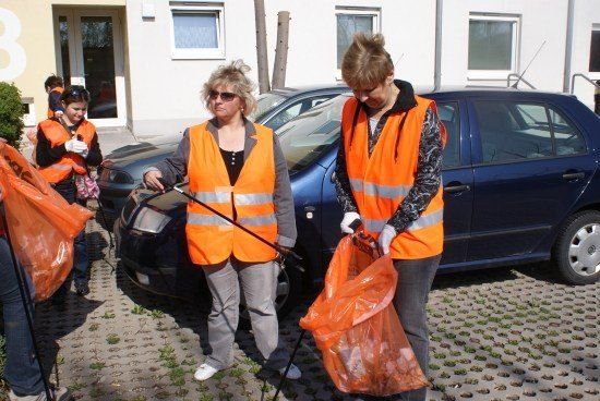 Zwei Frauen in orangenen Westen sammeln Müll vor einem Gebäude, in dessen Nähe Autos geparkt sind.