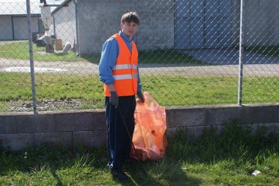 Ein Mann in einer orangefarbenen Sicherheitsweste und Handschuhen hält einen Plastikbeutel und steht neben einem Maschendrahtzaun und einer Betonmauer.