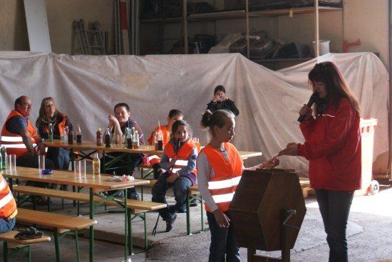 Eine Gruppe von Menschen in orangefarbenen Westen sitzt um Tische in einem Raum herum. Eine Frau in einer roten Jacke spricht in ein Mikrofon.