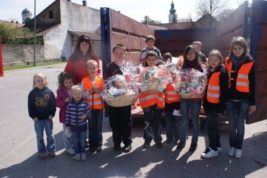 Eine Gruppe von Kindern und Erwachsenen steht vor einem Lastwagen und hält Körbe mit Gegenständen. Sie tragen orange Westen.