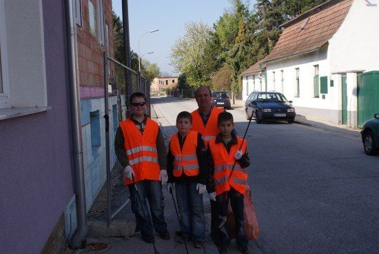 Vier Personen, darunter ein Mann und drei Jungen, stehen auf einer Straße und tragen orange Westen. Sie halten Stöcke in der Hand und scheinen den Bereich zu reinigen.