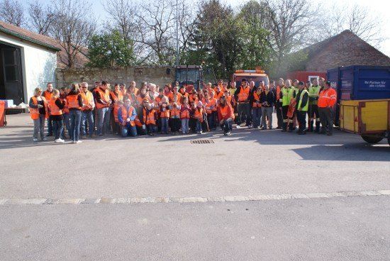 Eine Gruppe von Menschen in orangefarbenen Westen versammelt sich vor einem Gebäude mit geparkten Maschinenfahrzeugen dahinter.