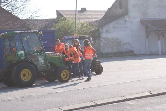 Eine Gruppe von Menschen in orangefarbenen Westen geht auf einer Straße, mit Traktoren und einem blauen Müllcontainer im Hintergrund.