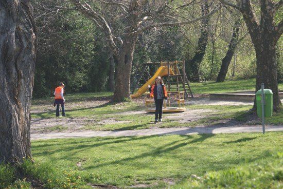 Zwei Personen in orange Westen gehen in einem Park mit einem gelben Rutschen und einem grünen Mülleimer.
