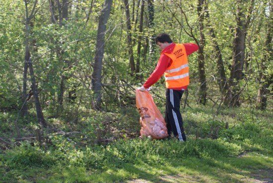 Ein Mann in einer orangefarbenen Warnweste sammelt Müll in einem Wald. Er hält einen orangefarbenen Beutel voller Abfall.