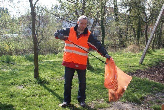 Ein Mann in einer orangefarbenen Warnweste hält einen langen Stock und einen orangefarbenen Müllsack, während er in einem Grasbereich steht.
