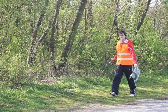 Ein Mann in einer orangefarbenen Warnweste geht auf einem Grasweg in der Nähe eines Waldes. Er hält ein Werkzeug und eine Tasche.