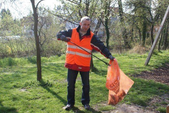 Ein Mann in einer orangefarbenen Weste hält einen Plastiksack in einer grasbewachsenen Fläche.